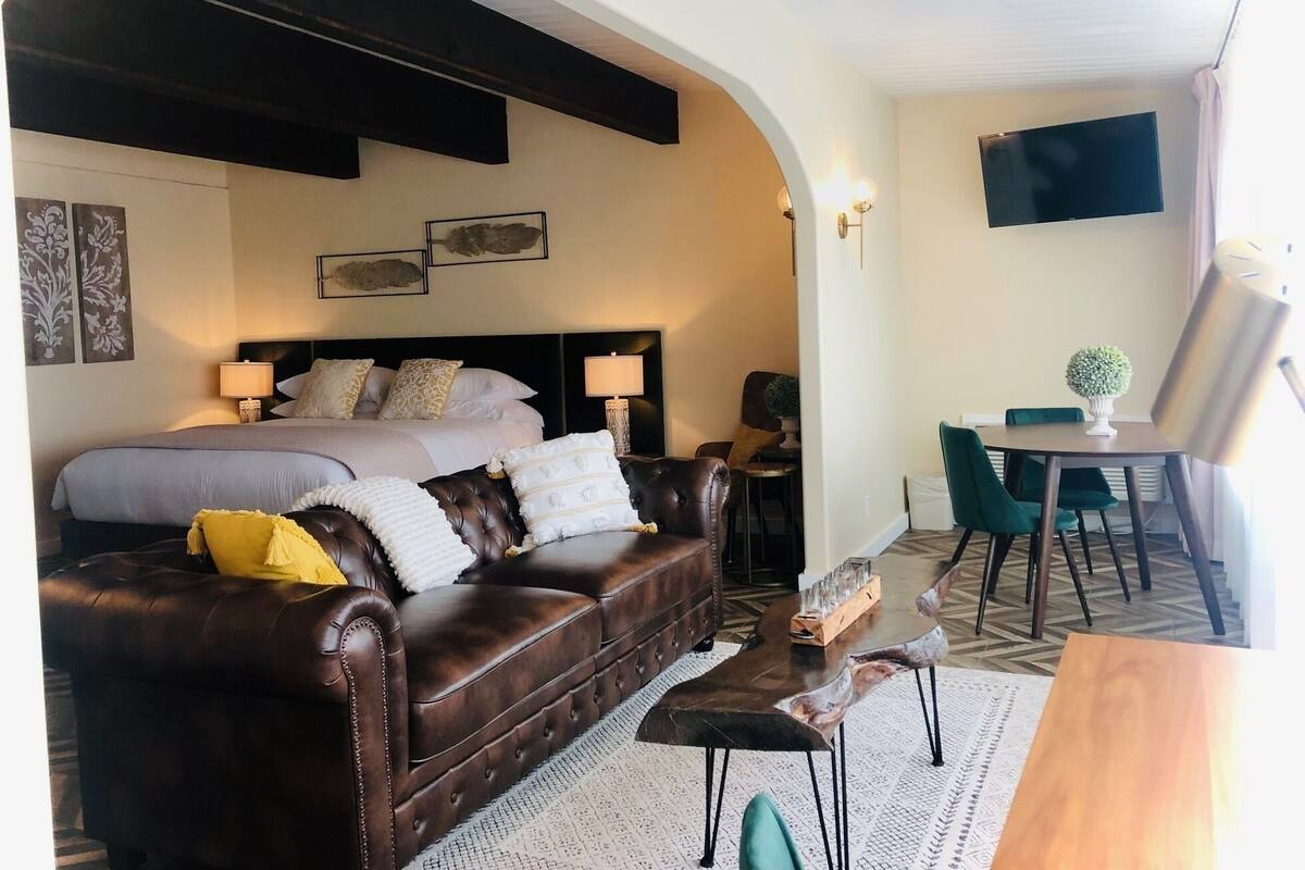 A hotel room with a large bed, a leather sofa, a dining table with chairs, and a TV mounted on the wall.