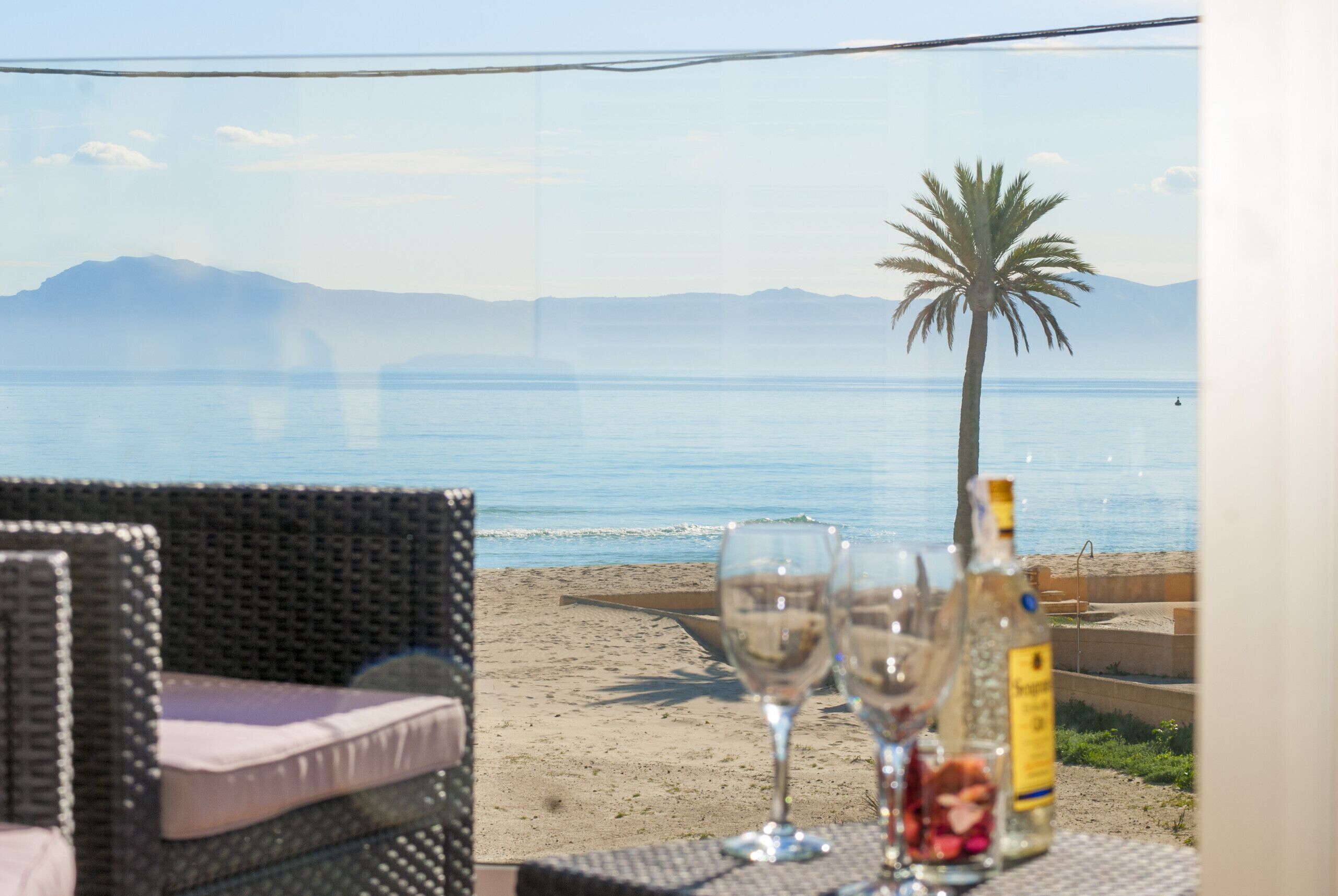 Een tafel aan het strand met twee wijnglazen en een fles, met uitzicht op een zanderig strand, een palmboom en een berg in de verte.