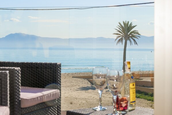 A beachside table with two wine glasses and a bottle, overlooking a sandy beach with a palm tree and a mountain in the distance.