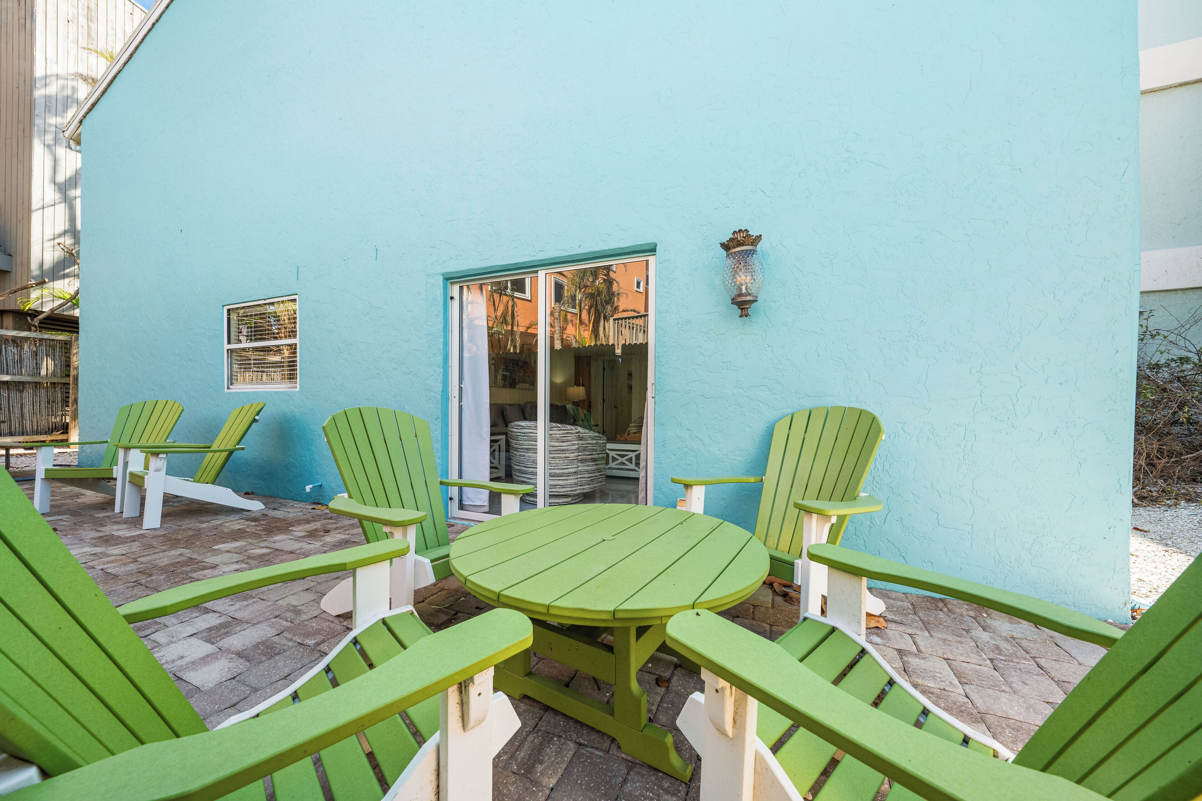 En terrasse med et rundt bord og seks grønne stole, en lyseblå væg og en dør ind til et rum indendørs.