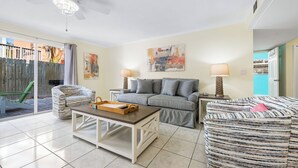 Una sala de estar con un sofá, una mesa de centro y una silla. Hay un ventanal corredero que da a un patio.