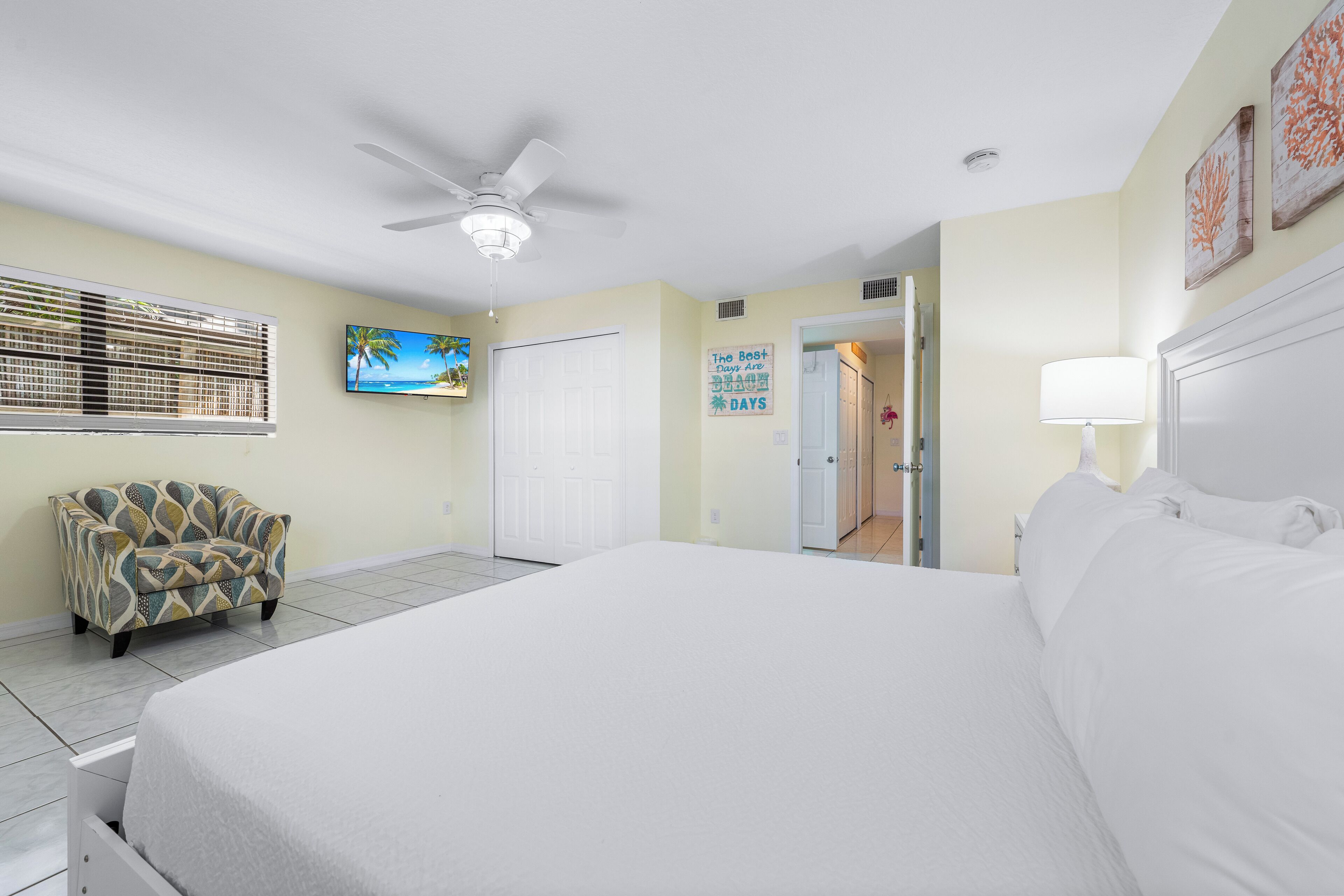 A bedroom with a large bed, a ceiling fan, a TV, and a door leading to another room.