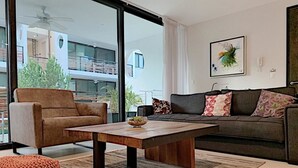 Uma sala de estar moderna com sofá, poltrona e um banquinho, uma mesa de centro de madeira, e vista para uma varanda com plantas.