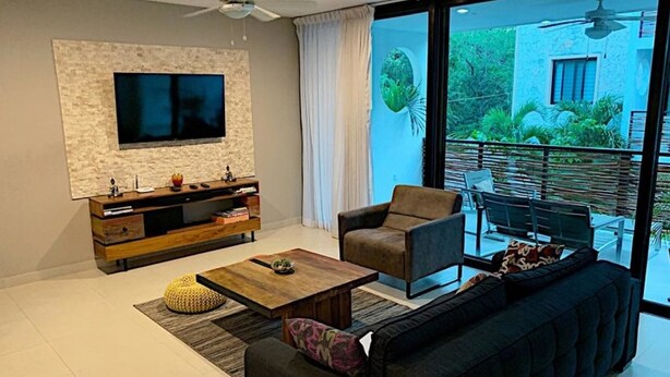 Sala de estar moderna com televisão de tela plana fixada em uma parede texturizada, móvel para home theater em madeira, sofá escuro, mesa de centro e uma grande porta de vidro deslizante que dá para um pátio externo com vista para o verde.