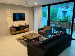 A modern living room with a flat-screen TV mounted on a textured wall, a wooden entertainment unit, a dark sofa, a coffee table, and a large sliding glass door leading to an outdoor patio with a view of greenery.