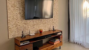 Uma sala de estar moderna com uma TV de tela plana fixada em uma parede texturizada, um móvel de entretenimento de madeira com livros e objetos decorativos, e uma janela grande com cortinas leves.