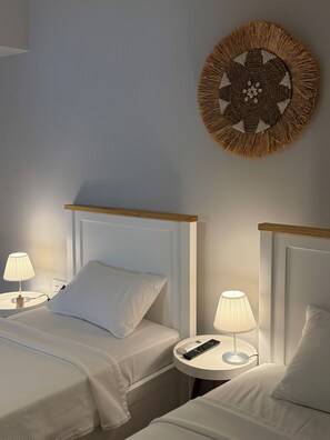 Habitación de hotel con dos camas, cada una con cabecero blanco y almohadas a juego. Hay dos mesitas de noche, una con una lámpara y un control remoto. Se observa un adorno en la pared sobre las camas.