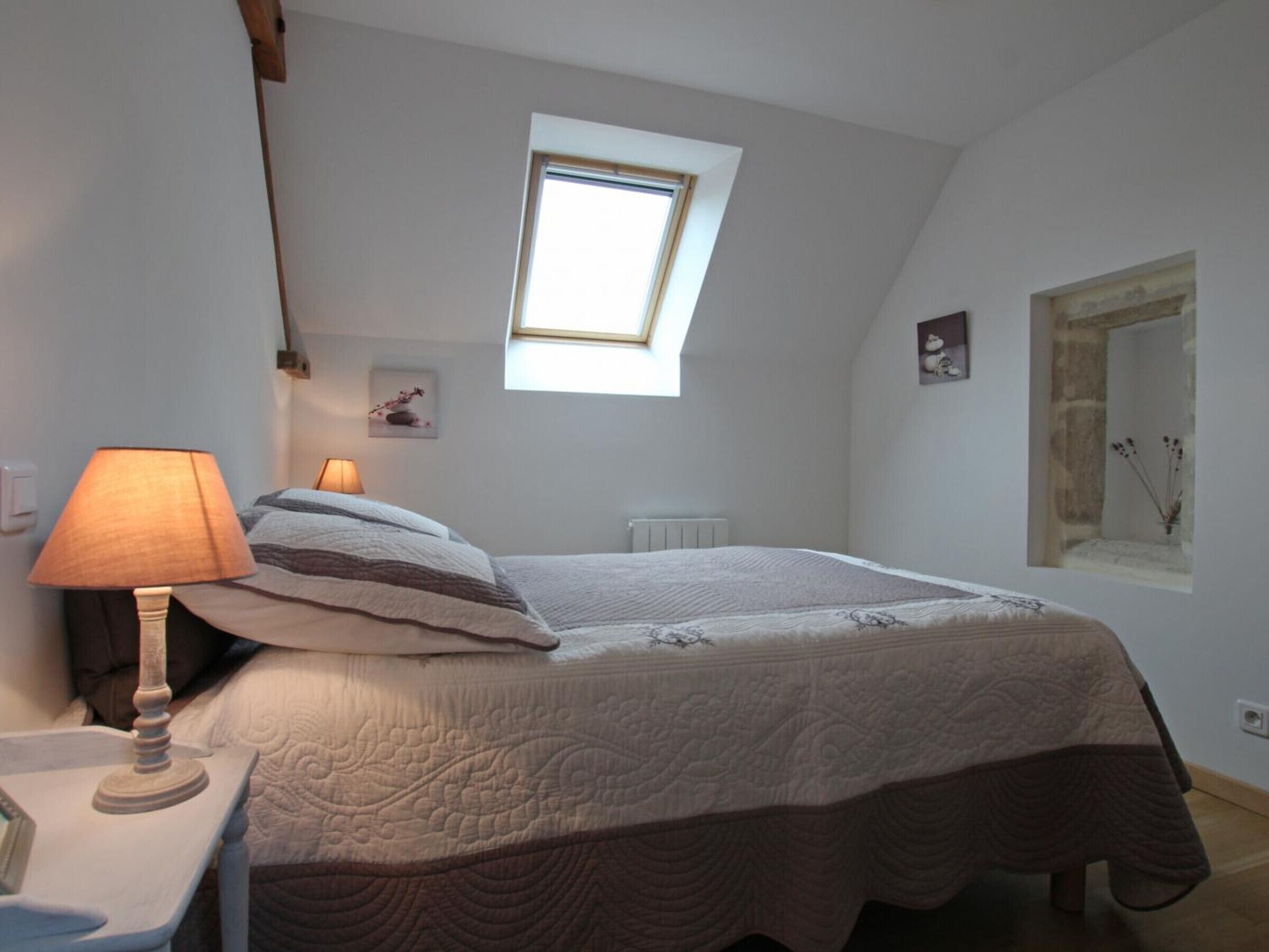 A bedroom with a skylight, a bed with a quilt, a bedside lamp, and a small picture on the wall.