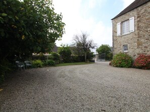 Une allée de gravier mène à un bâtiment en pierre doté d’une entrée fermée par un portail, le tout étant entouré de verdure et agrémenté d’un banc.