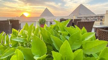 Terraço no telhado com plantas verdejantes, móveis de vime e vista para as pirâmides ao pôr do sol.