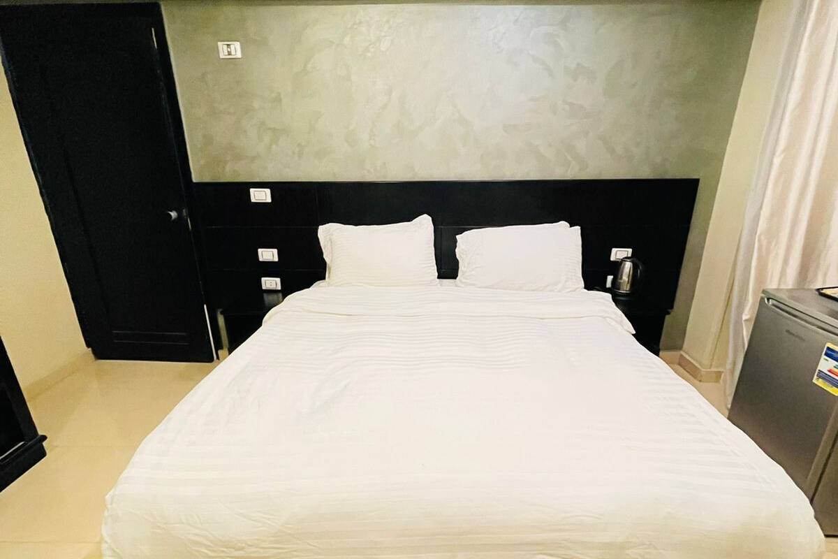 A hotel room with a large bed, a headboard with controls, a small refrigerator, and a door to the left.