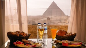Mesa de café da manhã com pão, queijo e bebidas, com vista para uma pirâmide.