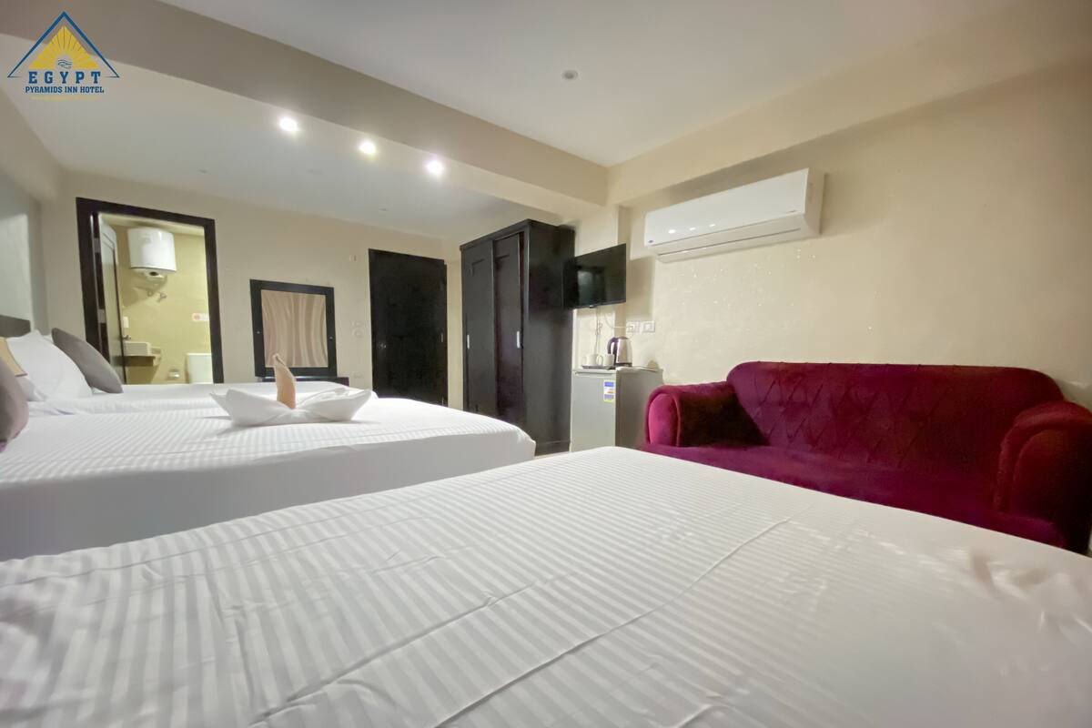 A hotel room with two beds, a red sofa, an air conditioning unit, and a television.