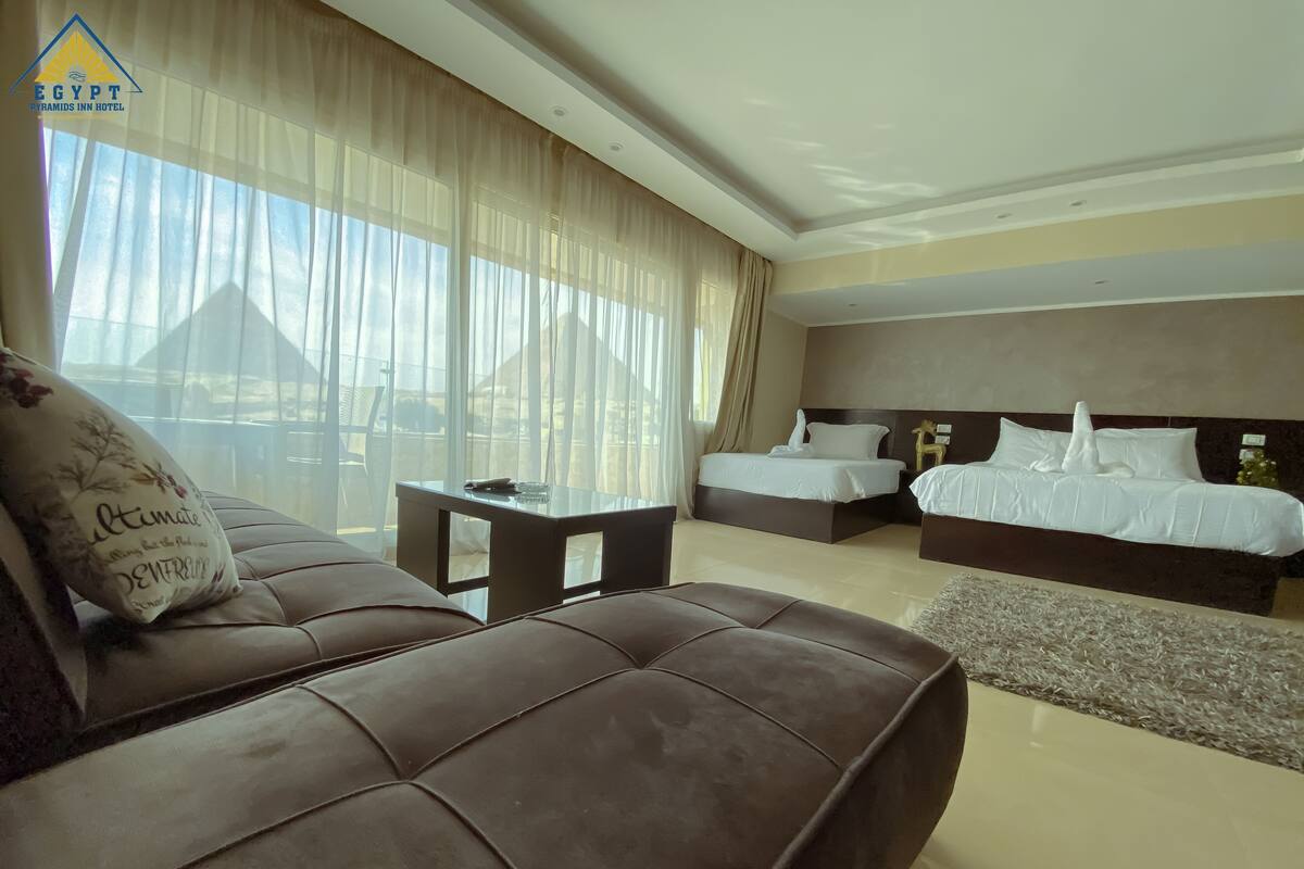 A hotel room with a brown leather sofa, a small table, two beds with white bedding, and sheer curtains.