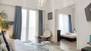 A modern living room with a glass coffee table, a white chair, a bed, and a balcony with blue curtains. - Philoxenia Flat (Athina)