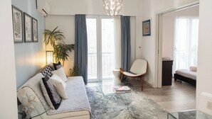 A modern living room with a sofa, a coffee table, a chair, and a large window with curtains. - Philoxenia Flat (Athina)