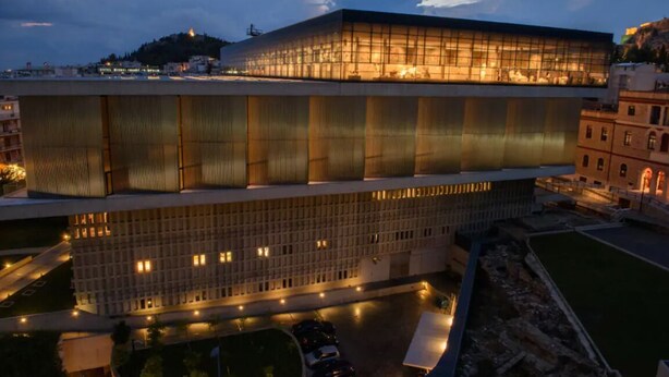 A modern building with a large glass facade and a lower section with a grid-like pattern. - Philoxenia Flat (Athina)