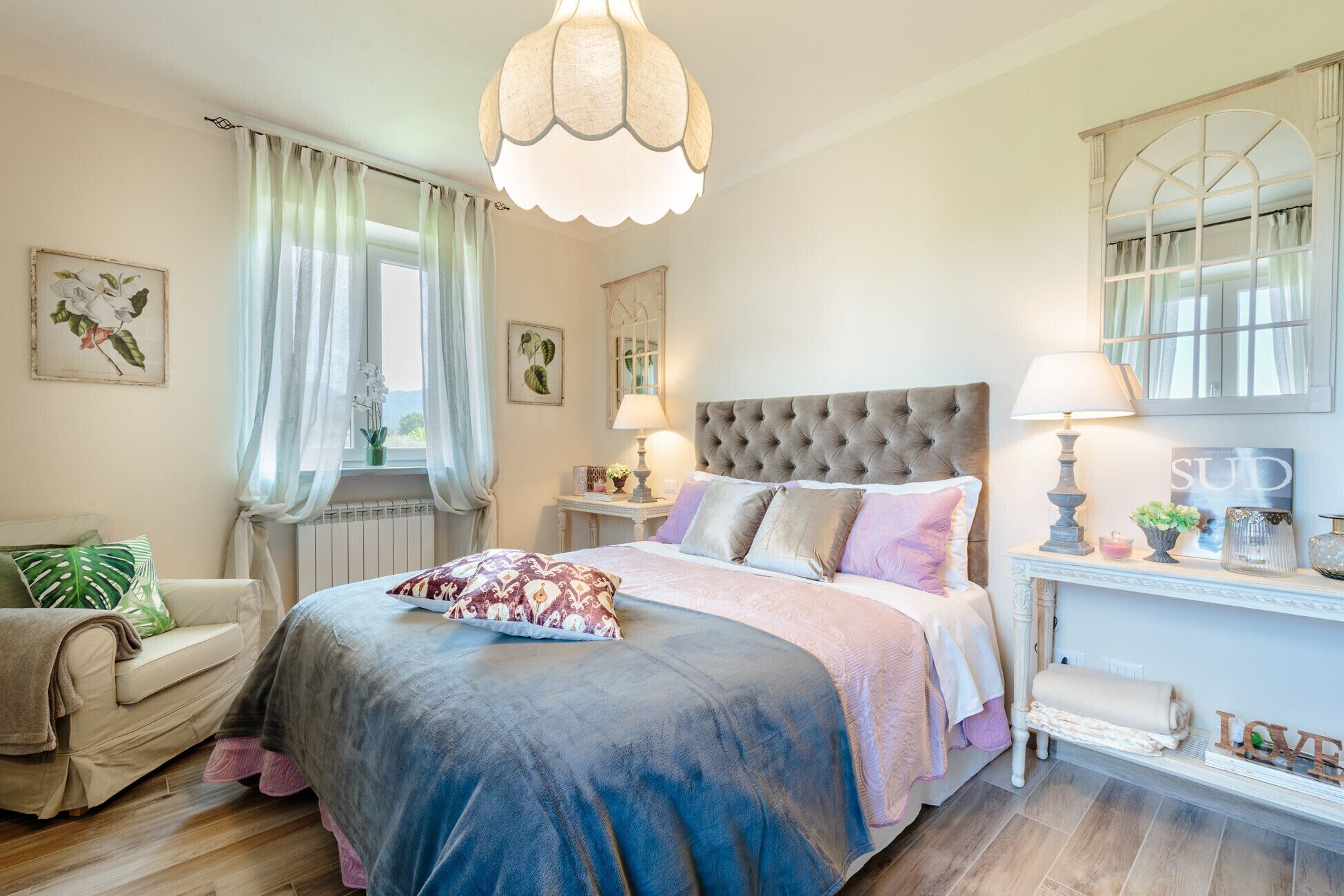 A bedroom with a large bed, two bedside tables, a chair, a window with curtains, and a chandelier.