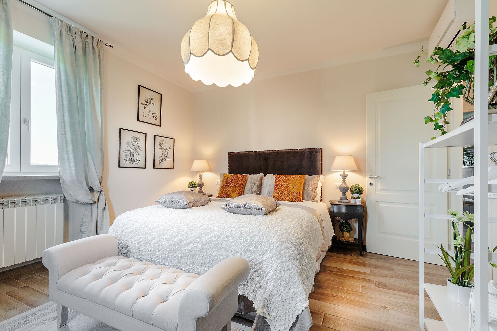 A bedroom with a large bed, two bedside tables with lamps, a white bench, a radiator, and a plant shelf.