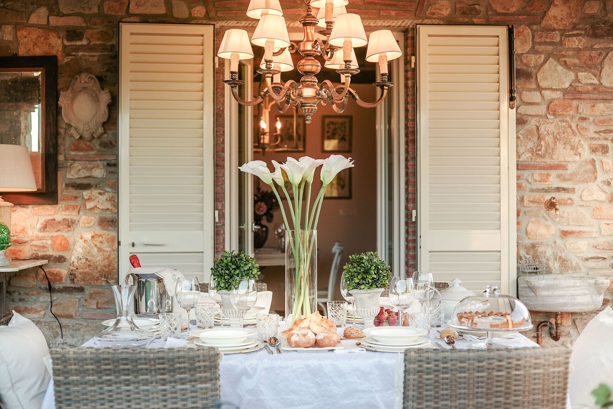 A dining table set for a meal with a chandelier above, a vase of flowers, and a stone wall in the background.
