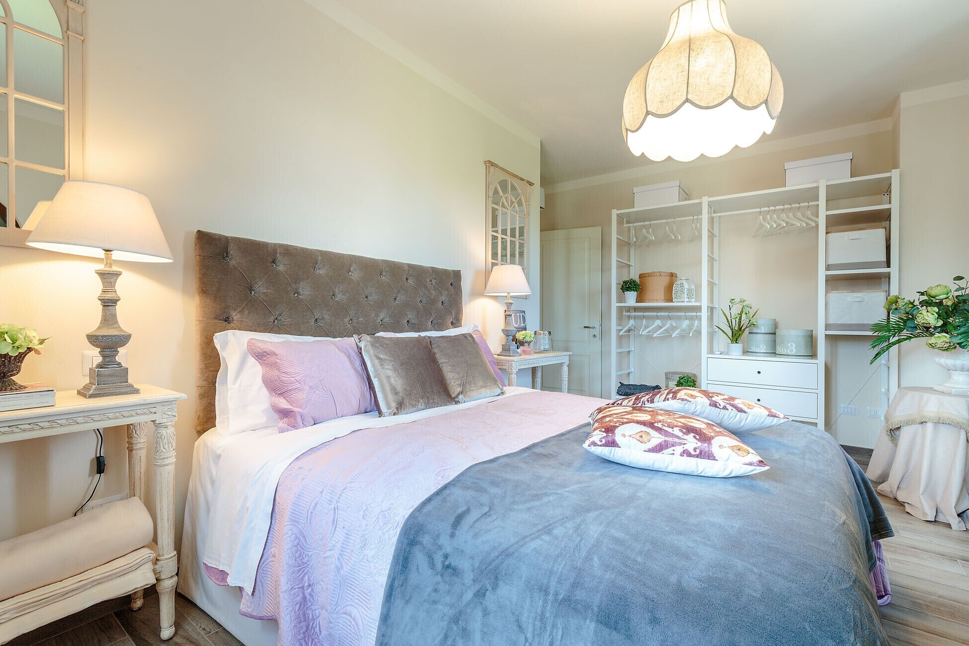 A bedroom with a tufted headboard, a large bed with multiple pillows, bedside lamps, a wardrobe with hanging space, and a decorative hanging light fixture.