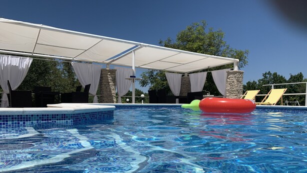 Une piscine dont le bord est recouvert de carreaux, un espace abrité avec des rideaux blancs et un flotteur gonflable rouge.