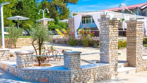 Un espace extérieur aménagé avec des sièges en pierre et des parasols, entouré de verdure et bordé d’un bâtiment au toit rouge.