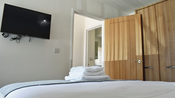 Quarto com cama, televisão fixada na parede, guarda-roupa de madeira e porta para outro espaço.