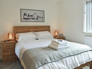 A neatly made bed with white linens, a grey checkered throw, and a folded towel on the footboard. There are two bedside tables with lamps, a framed artwork above the bed, and a window with blinds on the right.