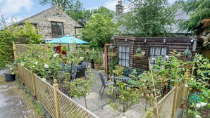 A garden with a wooden cabin, stone house, and a fenced patio area with outdoor seating.