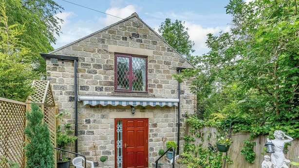 A stone house with a red door, a small porch, and a garden with potted plants and a statue.