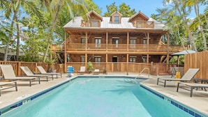 A pool with lounge chairs, a wooden building with balconies, and tropical trees.