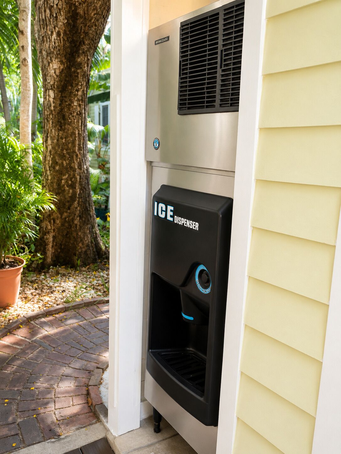 An ice dispenser attached to a building exterior, with a tree and potted plant nearby.