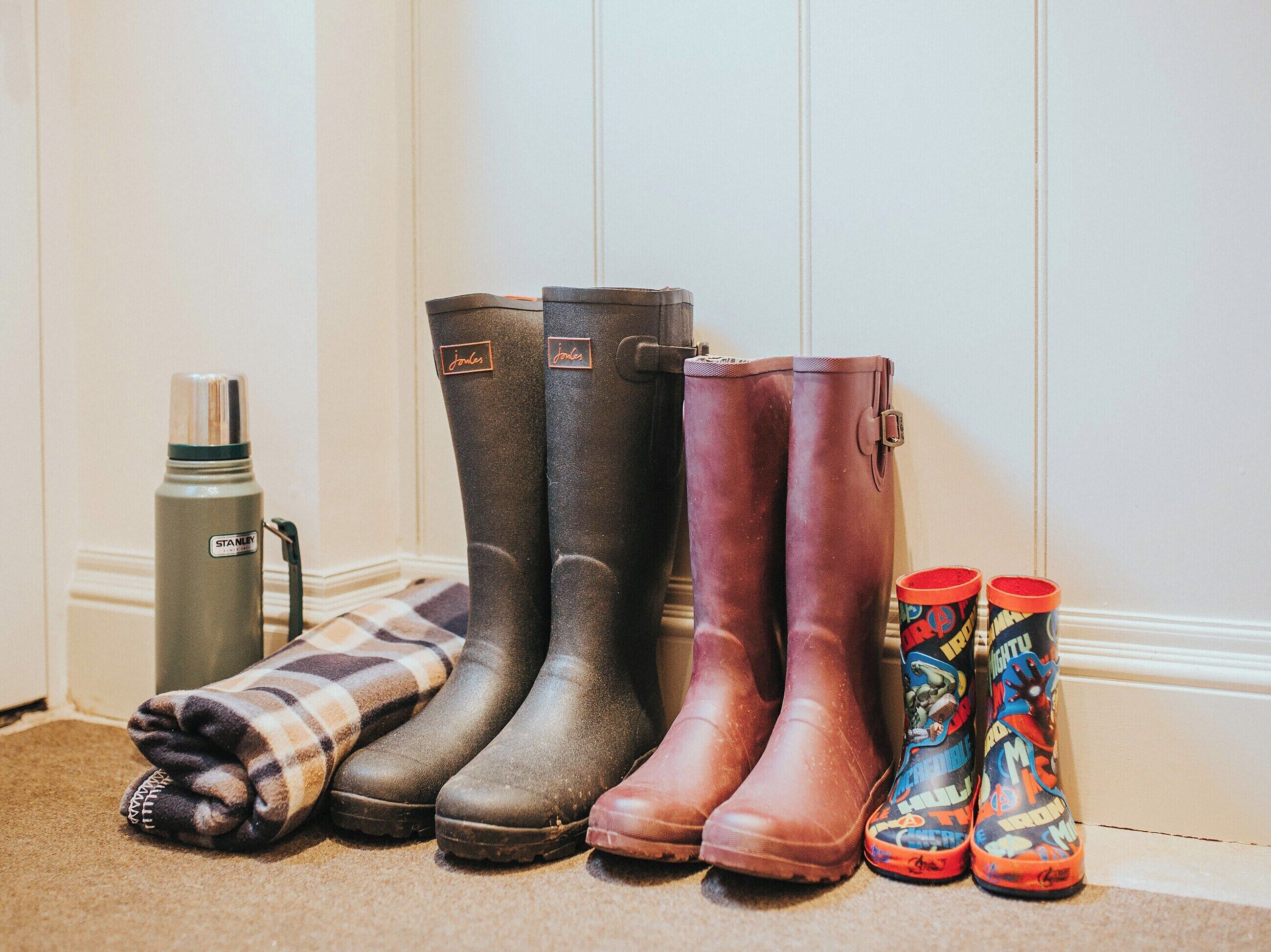 A row of boots and a thermos on a carpeted floor against a white wall.