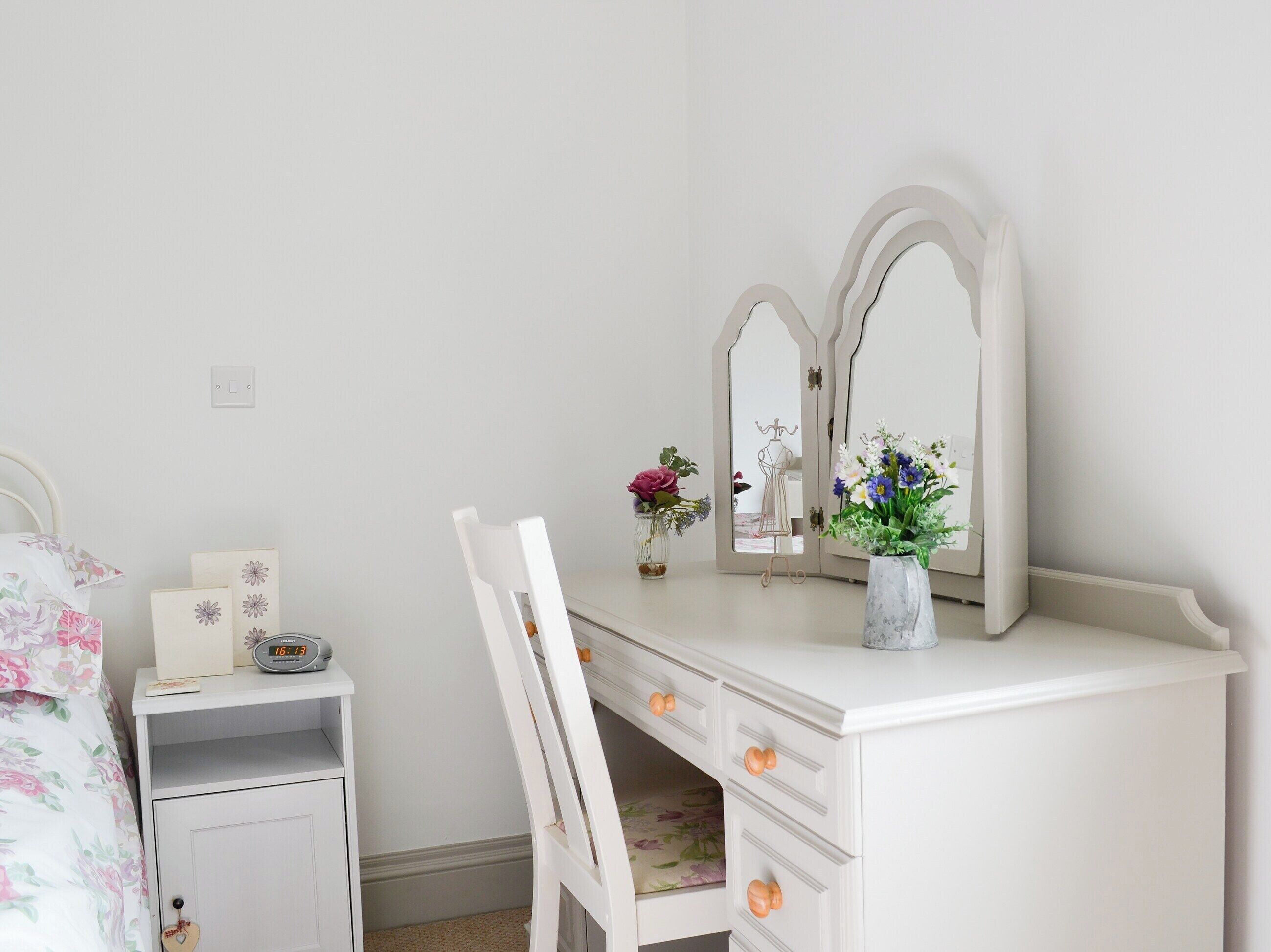 Une coiffeuse blanche avec deux miroirs, une chaise, une petite table avec une horloge et un vase rempli de fleurs.