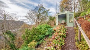 A green wooden structure with a bench and a table, surrounded by a variety of plants and trees. - 4 bedroom accommodation in Plockton (Plockton)