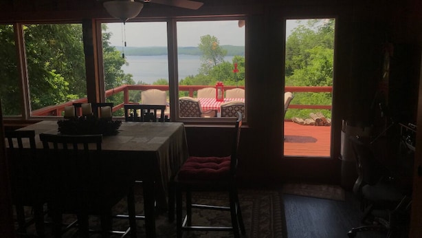A dining area with a table and chairs, a ceiling fan, and a view of a lake and greenery through large windows. - Amazing view of Lake Norfork, room for everyone! (Mountain Home)