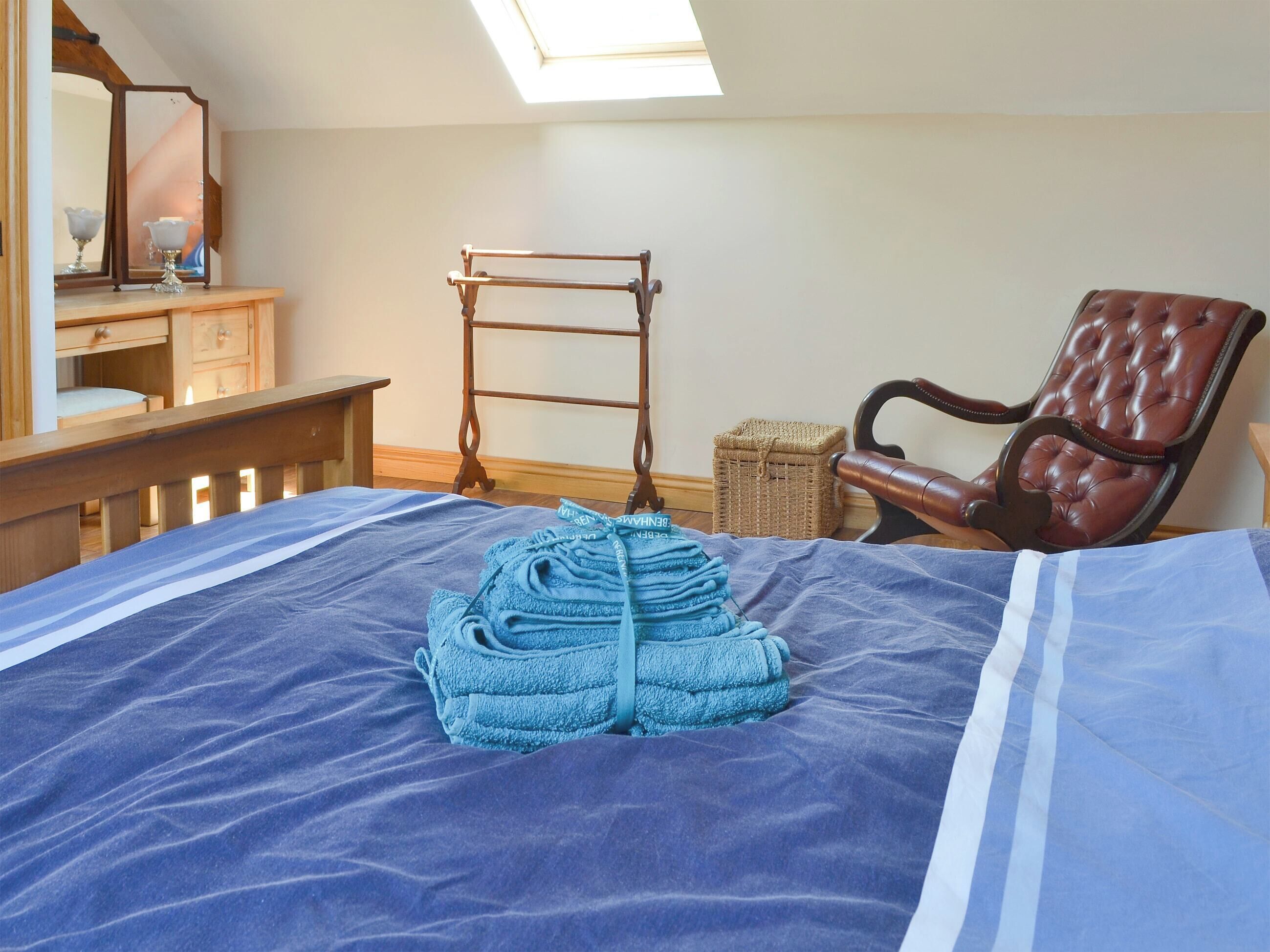 A neatly made bed with a blue blanket and a stack of blue towels. There is a wooden chair and a small wooden table with a lamp. A wooden ladder is leaning against the wall.