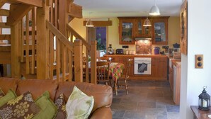 Un soggiorno accogliente con un divano in pelle marrone, una scala in legno e un angolo cucina con mobili in legno e zoccolo in pietra.