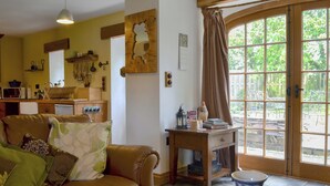 Un accogliente soggiorno con un divano in pelle, un tavolino in legno e un vaso. È presente un angolo cottura con utensili a parete, una finestra con tende e vista su del verde all'esterno.