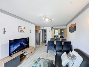 A modern living room with a flat-screen TV, a glass coffee table, a black leather sofa, and a dining area with a wooden cabinet and chairs.