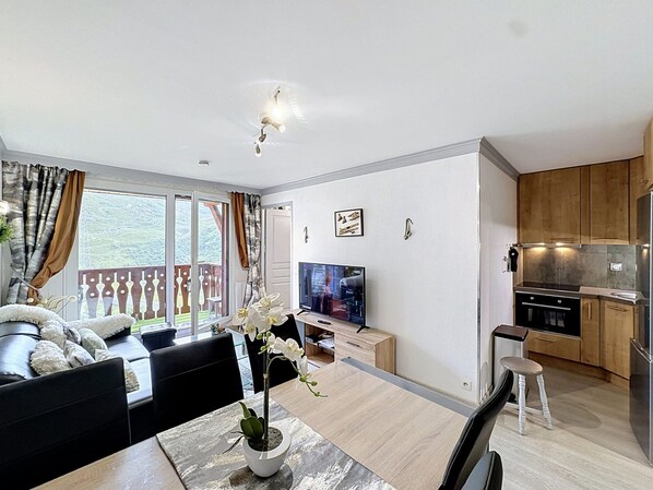 Una sala de estar moderna con un sofá, una mesa de comedor y un televisor. Hay un balcón con vistas al exterior.