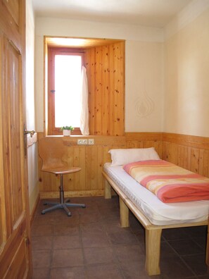 Een kleine kamer met een houten bed, een tafeltje, een raam met gordijn en een deur.