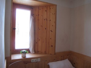 Ein Zimmer mit Holzwänden, einem Fenster mit Vorhang, einer kleinen Pflanze auf einem Sims und einem Bett mit Kopfkissen.