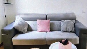 A grey sofa with two pink and two grey cushions, a small round table with a white object, and a lamp on a shelf.
