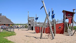 Une plage dotée d’une aire de jeux en forme de bateau de pirates en bois, comprenant une roue de gouvernail et une tour d’observation.