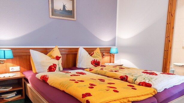 A double bed with floral bedding, a wooden headboard, bedside tables with lamps, and a framed picture on the wall. - Double room 4 - Rankwitzer Hof (Rankwitz)