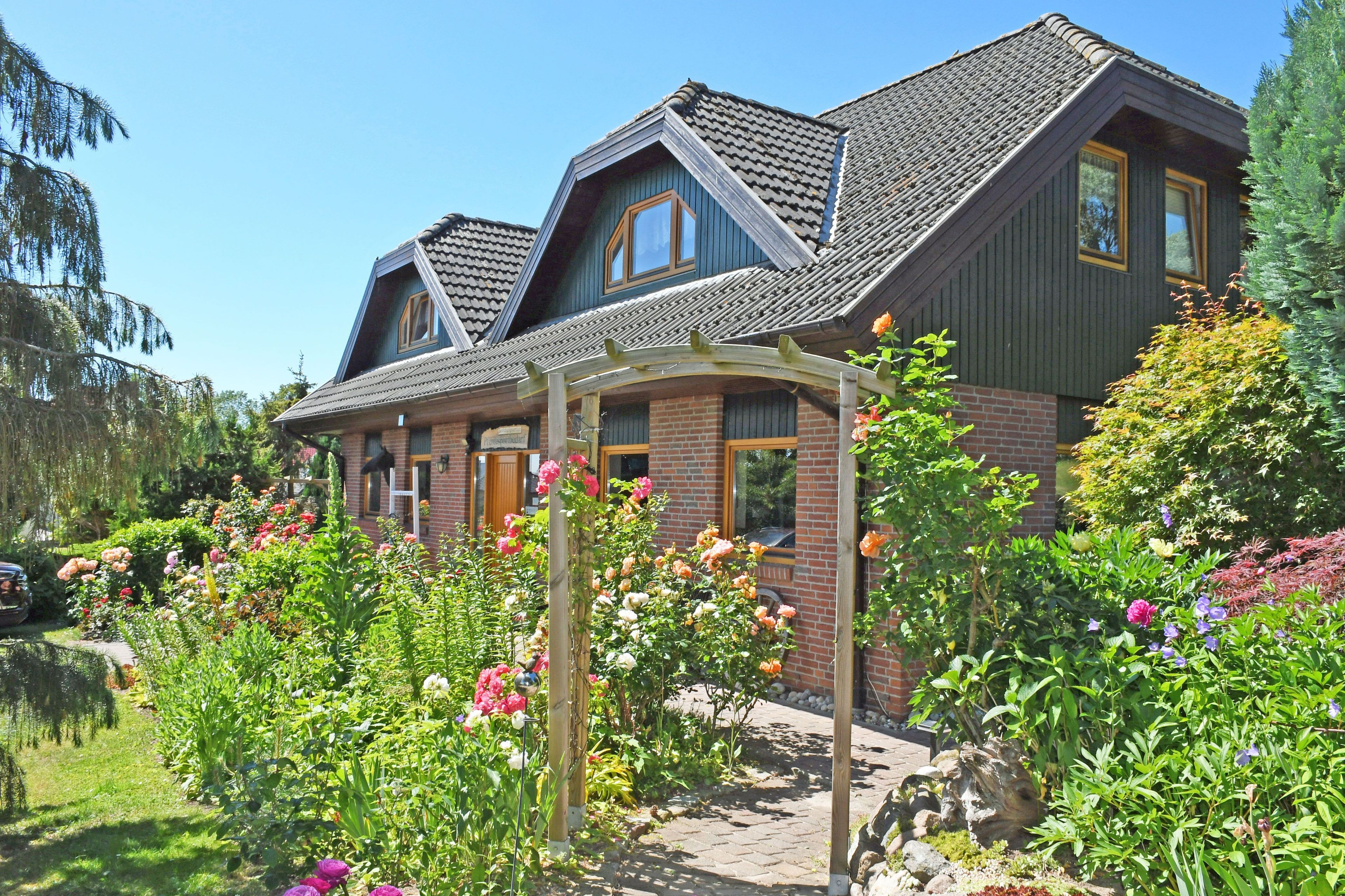 A two-story house with a brick facade, a sloped roof, and a garden with various flowers.