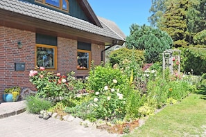 Et murstenshus med tegltag, en have med forskellige planter og blomster, samt en træpergola med blomster.