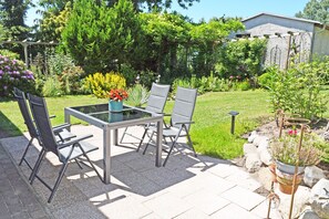 Une terrasse extérieure avec une table en verre et quatre chaises, entourée d’un jardin où poussent différentes plantes et fleurs.